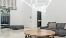 Ruang keluarga yang tertata rapi dengan banyak cahaya alami dan sentuhan tanaman hijau, mencerminkan prinsip-prinsip feng shui modern untuk menciptakan suasana yang harmonis dan menenangkan. Penataan ini bertujuan untuk memperlancar aliran energi positif di dalam rumah.